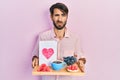 Young hispanic man holding tray with breakfast food and heart draw clueless and confused expression Royalty Free Stock Photo