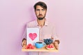 Young hispanic man holding tray with breakfast food and heart draw puffing cheeks with funny face Royalty Free Stock Photo