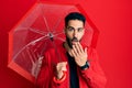 Young hispanic man holding umbrella covering mouth with hand, shocked and afraid for mistake