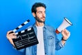 Young hispanic man holding video film clapboard and megaphone smiling looking to the side and staring away thinking