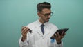 Young hispanic man in lab coat holds metallic engine component and reads digital tablet in teal studio; focus Royalty Free Stock Photo