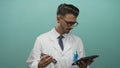 Young hispanic man in lab coat holds metallic engine component and reads digital tablet in teal studio; focus Royalty Free Stock Photo