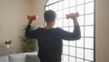 A young hispanic man lifting dumbbells in an interior home setting with natural light filtering through a large window, showcasing Royalty Free Stock Photo