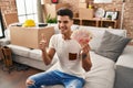 Young hispanic man moving to a new home holding shekels smiling happy and positive, thumb up doing excellent and approval sign Royalty Free Stock Photo