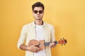 Young hispanic man playing ukulele over yellow background puffing cheeks with funny face