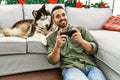 Young hispanic man playing video game sitting on sofa with dog by christmas decor at home Royalty Free Stock Photo