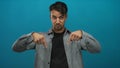 Young hispanic man pointing down against a blue background, emphasizing direction and focus in a casual setting, ideal for Royalty Free Stock Photo