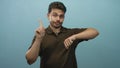 Young hispanic man pointing finger and checking wrist while looking at wrist in light blue studio; time pressure impatience Royalty Free Stock Photo