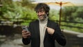 Young hispanic man pointing at mobile while smiling outdoors in park setting, capturing a joyful and engaging moment in natural Royalty Free Stock Photo