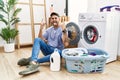 Young hispanic man putting dirty laundry into washing machine shouting with crazy expression doing rock symbol with hands up Royalty Free Stock Photo