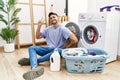 Young hispanic man putting dirty laundry into washing machine smiling looking to the camera showing fingers doing victory sign Royalty Free Stock Photo