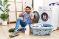 Young hispanic man putting dirty laundry into washing machine trying to hear both hands on ear gesture, curious for gossip