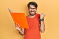 Young hispanic man reading book screaming proud, celebrating victory and success very excited with raised arm