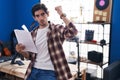 Young hispanic man reading music sheet at music studio annoyed and frustrated shouting with anger, yelling crazy with anger and Royalty Free Stock Photo