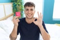 Young hispanic man sitting on the bed holding condom doing ok sign with fingers, smiling friendly gesturing excellent symbol Royalty Free Stock Photo