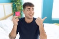 Young hispanic man sitting on the bed holding condom pointing thumb up to the side smiling happy with open mouth Royalty Free Stock Photo