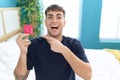 Young hispanic man sitting on the bed holding condom smiling happy pointing with hand and finger Royalty Free Stock Photo