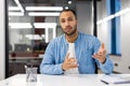 Young hispanic man sitting in the office at the desk, looking and talking to the camera while gesturing remotely with Royalty Free Stock Photo