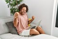 Young hispanic man sitting on the sofa at home using laptop very happy and excited doing winner gesture with arms raised, smiling Royalty Free Stock Photo