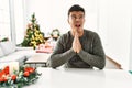 Young hispanic man sitting on the table by christmas tree begging and praying with hands together with hope expression on face Royalty Free Stock Photo