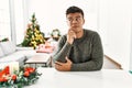 Young hispanic man sitting on the table by christmas tree with hand on chin thinking about question, pensive expression Royalty Free Stock Photo