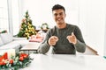 Young hispanic man sitting on the table by christmas tree pointing fingers to camera with happy and funny face Royalty Free Stock Photo