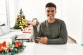 Young hispanic man sitting on the table by christmas tree smiling cheerful presenting and pointing with palm of hand looking at Royalty Free Stock Photo