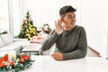 Young hispanic man sitting on the table by christmas tree smiling with hand over ear listening an hearing to rumor or gossip