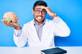 Young hispanic man sitting at the table wearing doctor coat holding brain stressed and frustrated with hand on head, surprised and Royalty Free Stock Photo