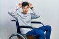 Young hispanic man sitting on wheelchair doing funny gesture with finger over head as bull horns