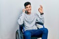 Young hispanic man sitting on wheelchair talking on the smartphone doing ok sign with fingers, smiling friendly gesturing Royalty Free Stock Photo
