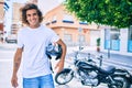 Young hispanic man smiling happy holding moto helmet over motorcycle at the city