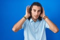 Young hispanic man standing over blue background trying to hear both hands on ear gesture, curious for gossip Royalty Free Stock Photo