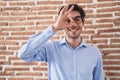 Young hispanic man standing over brick wall background doing ok gesture with hand smiling, eye looking through fingers with happy Royalty Free Stock Photo