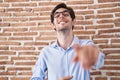 Young hispanic man standing over brick wall background laughing at you, pointing finger to the camera with hand over body, shame