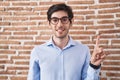 Young hispanic man standing over brick wall background showing and pointing up with finger number one while smiling confident and Royalty Free Stock Photo