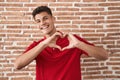 Young hispanic man standing over bricks wall smiling in love doing heart symbol shape with hands