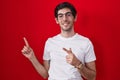 Young hispanic man standing over red background smiling and looking at the camera pointing with two hands and fingers to the side Royalty Free Stock Photo