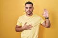 Young hispanic man standing over yellow background swearing with hand on chest and open palm, making a loyalty promise oath Royalty Free Stock Photo