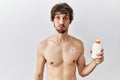 Young hispanic man standing shirtless holding sunscreen lotion puffing cheeks with funny face