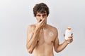 Young hispanic man standing shirtless holding sunscreen lotion smelling something stinky and disgusting, intolerable smell,