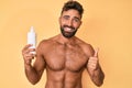 Young hispanic man standing shirtless holding sunscreen lotion smiling happy and positive, thumb up doing excellent and approval Royalty Free Stock Photo