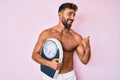Young hispanic man standing shirtless holding weighing machine pointing thumb up to the side smiling happy with open mouth Royalty Free Stock Photo