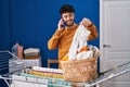 Young hispanic man talking on smartphone hanging clothes on clothesline at laundry room