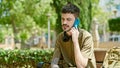 Young hispanic man talking on smartphone sitting on bench at park Royalty Free Stock Photo