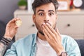 Young hispanic man with tattoos holding cardano cryptocurrency coin covering mouth with hand, shocked and afraid for mistake