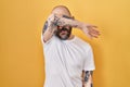 Young hispanic man with tattoos standing over yellow background covering eyes with arm, looking serious and sad Royalty Free Stock Photo