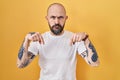 Young hispanic man with tattoos standing over yellow background pointing down looking sad and upset, indicating direction with Royalty Free Stock Photo