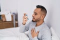 Young hispanic man using inhaler sitting on bed at bedroom Royalty Free Stock Photo
