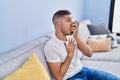 Young hispanic man using inhaler sitting on sofa at home